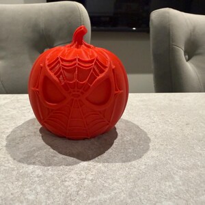 May include: A bright red, round pumpkin with a carved Spider-Man design. The pumpkin features the character's mask and web pattern. The pumpkin is sitting on a gray surface, with a shadow cast to the left. The theme is Halloween.