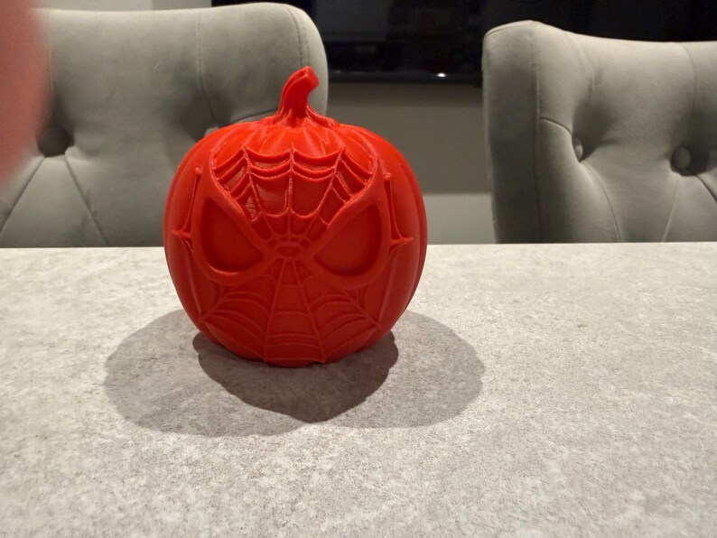 May include: A bright red, round pumpkin with a carved Spider-Man design. The pumpkin features the character's mask and web pattern. The pumpkin is sitting on a gray surface, with a shadow cast to the left. The theme is Halloween.