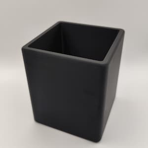 May include: A black, square-shaped container with a matte finish. The container has a rectangular opening and is designed with straight sides. The object is likely a desk organizer or a decorative storage item.