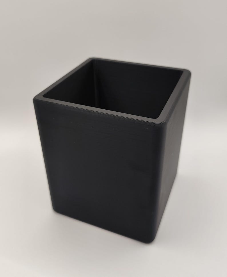 May include: A black, square-shaped container with a matte finish. The container has a rectangular opening and is designed with straight sides. The object is likely a desk organizer or a decorative storage item.