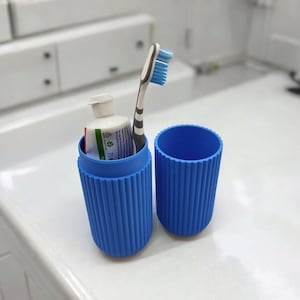 May include: A blue travel toothbrush holder with a matching lid. Inside the holder is a tube of toothpaste and a toothbrush with blue bristles. The holder is ribbed and cylindrical, designed for travel.
