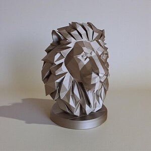 May include: A metallic silver lion head sculpture with a geometric, polygonal design. The lion's mane and face are intricately detailed, and the sculpture sits on a circular base. The artwork is a decorative item.