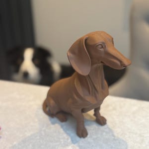 May include: A brown dachshund figurine, sitting upright with its ears flopped down. The dog is a solid color and appears to be made of a hard material. A black and white dog is visible in the background.
