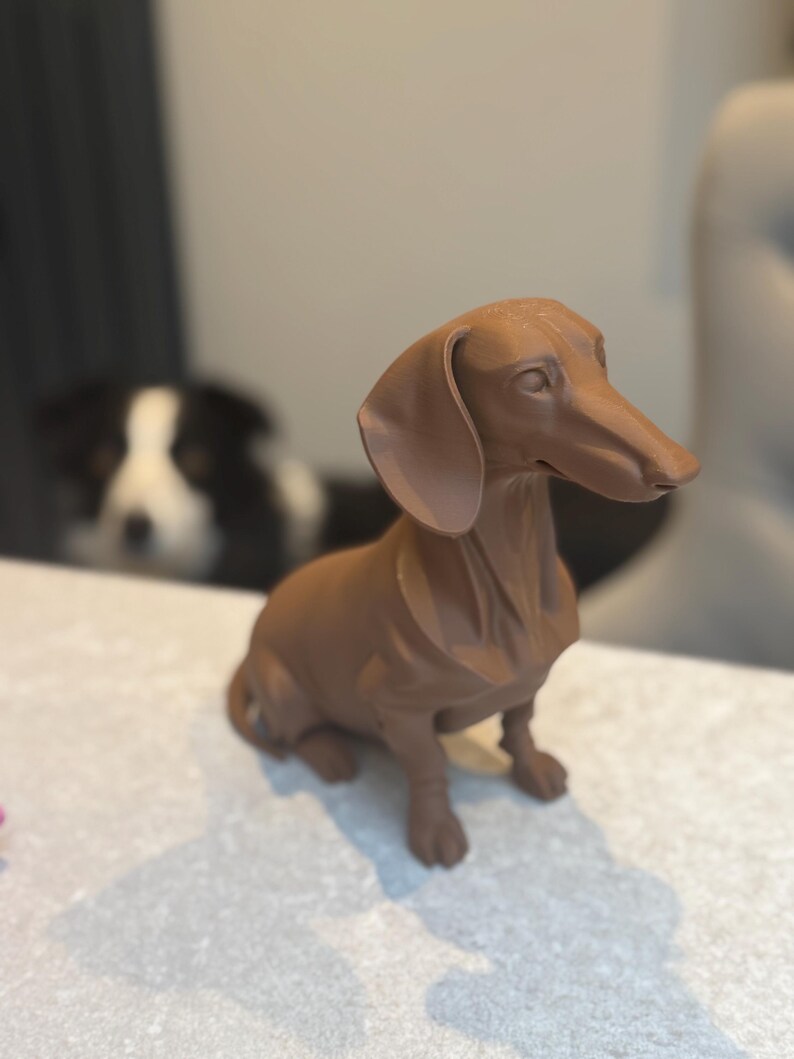 May include: A brown dachshund figurine, sitting upright with its ears flopped down. The dog is a solid color and appears to be made of a hard material. A black and white dog is visible in the background.