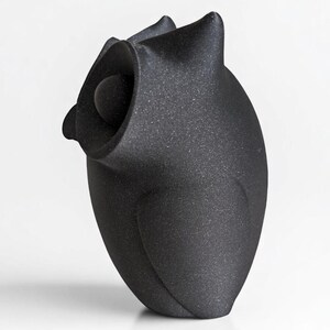 Op de afbeelding: Een zwarte, getextureerde uil sculptuur met een afgerond lichaam en een groot, open oog. De uil heeft puntige oorpluimen en een gladde, bolvormige kop. De sculptuur staat tegen een witte achtergrond.