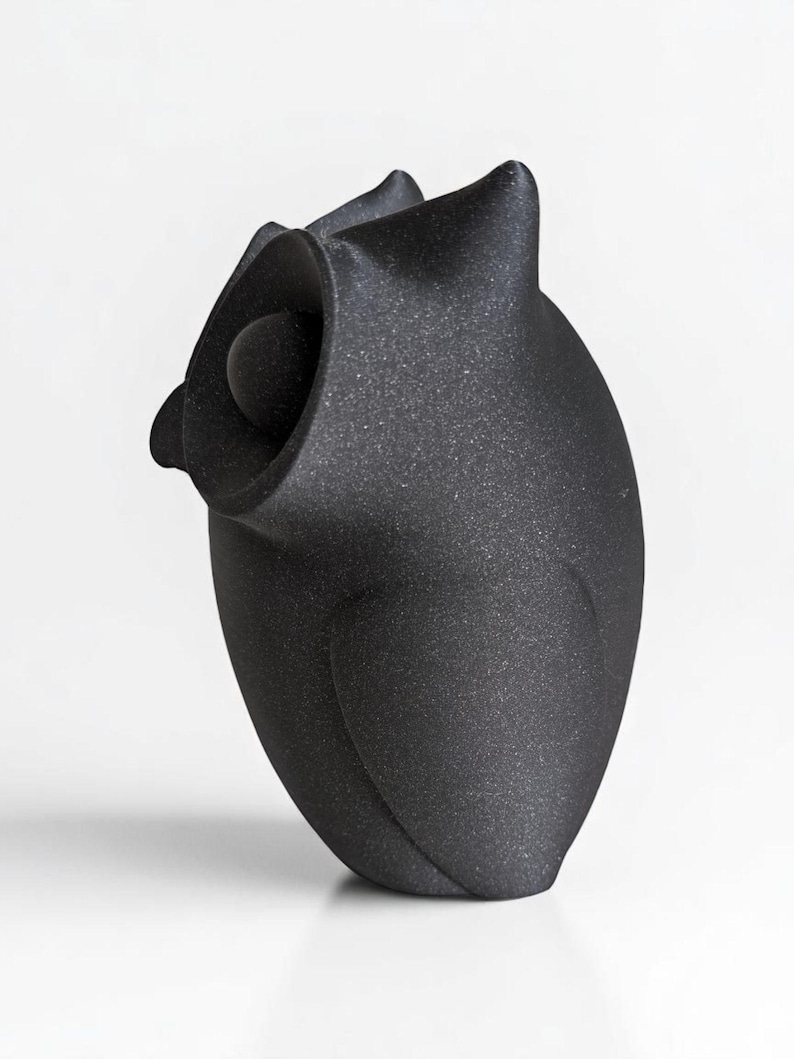 Op de afbeelding: Een zwarte, getextureerde uil sculptuur met een afgerond lichaam en een groot, open oog. De uil heeft puntige oorpluimen en een gladde, bolvormige kop. De sculptuur staat tegen een witte achtergrond.