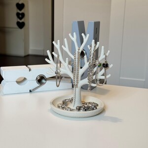 May include: A white jewelry tree with a tree branch design, holding several silver necklaces and bracelets. The base of the tree has a small dish with more jewelry. A large white clothespin is in the background.
