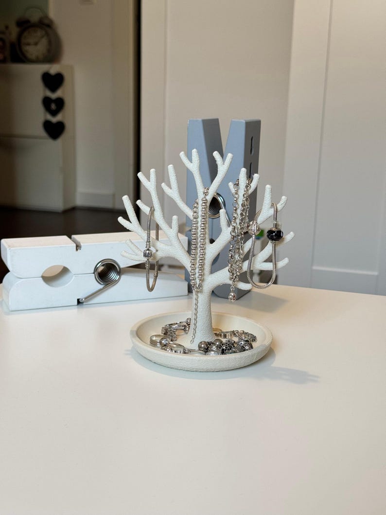 May include: A white jewelry tree with a tree branch design, holding several silver necklaces and bracelets. The base of the tree has a small dish with more jewelry. A large white clothespin is in the background.