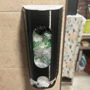 May include: A black plastic bag dispenser mounted on a beige tiled wall. The dispenser has a rectangular shape with a large oval opening revealing plastic bags inside. A white rim tops the dispenser, and a bag is visible at the bottom.