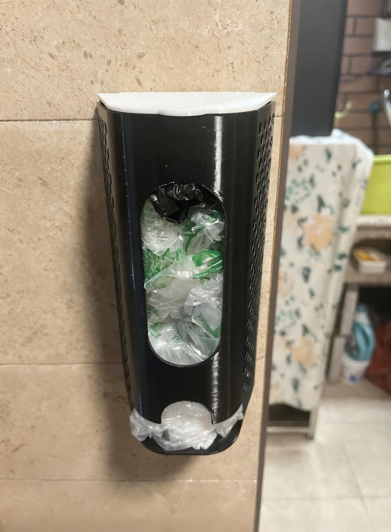 May include: A black plastic bag dispenser mounted on a beige tiled wall. The dispenser has a rectangular shape with a large oval opening revealing plastic bags inside. A white rim tops the dispenser, and a bag is visible at the bottom.
