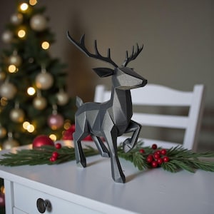 May include: A geometric, dark gray reindeer figurine with detailed antlers, posed in a running stance. The decorative piece is placed on a white surface with Christmas decorations in the background, including a Christmas tree and ornaments.