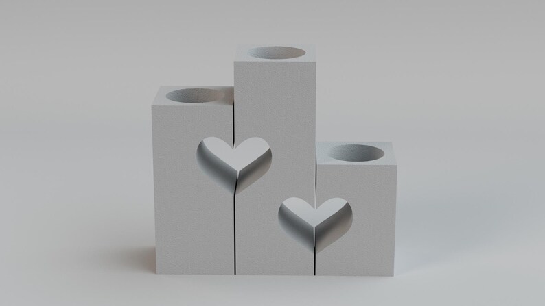 May include: A gray, tiered candle holder with heart cutouts. The holder has three sections of varying heights, each with a circular opening at the top for candles. The design features two heart-shaped cutouts.