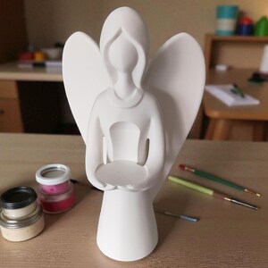 May include: A white angel figurine with outstretched wings and a circular platform held in its hands. The angel is standing on a wooden surface, with paint jars and paintbrushes nearby. The figurine is likely for decorative purposes.