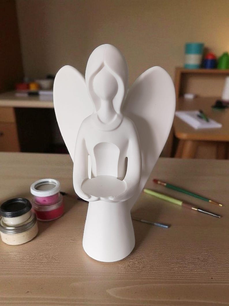 May include: A white angel figurine with outstretched wings and a circular platform held in its hands. The angel is standing on a wooden surface, with paint jars and paintbrushes nearby. The figurine is likely for decorative purposes.