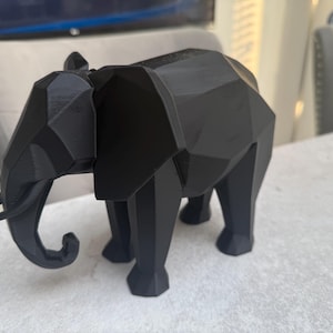 Op de afbeelding: Een zwarte geometrische olifant sculptuur. De olifant heeft een facetontwerp, met scherpe hoeken en vlakke oppervlakken. De slurf is gekruld en de slagtanden zijn zichtbaar. De sculptuur staat op een lichtgekleurd oppervlak.