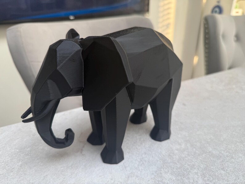 Op de afbeelding: Een zwarte geometrische olifant sculptuur. De olifant heeft een facetontwerp, met scherpe hoeken en vlakke oppervlakken. De slurf is gekruld en de slagtanden zijn zichtbaar. De sculptuur staat op een lichtgekleurd oppervlak.