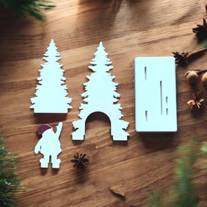 May include: White Christmas-themed cutouts on a wooden surface. The cutouts include two Christmas trees, a Santa Claus figure, and a rectangular piece with vertical slits. The Santa figure is wearing a red hat. The scene is decorated with pine needles and star anise.