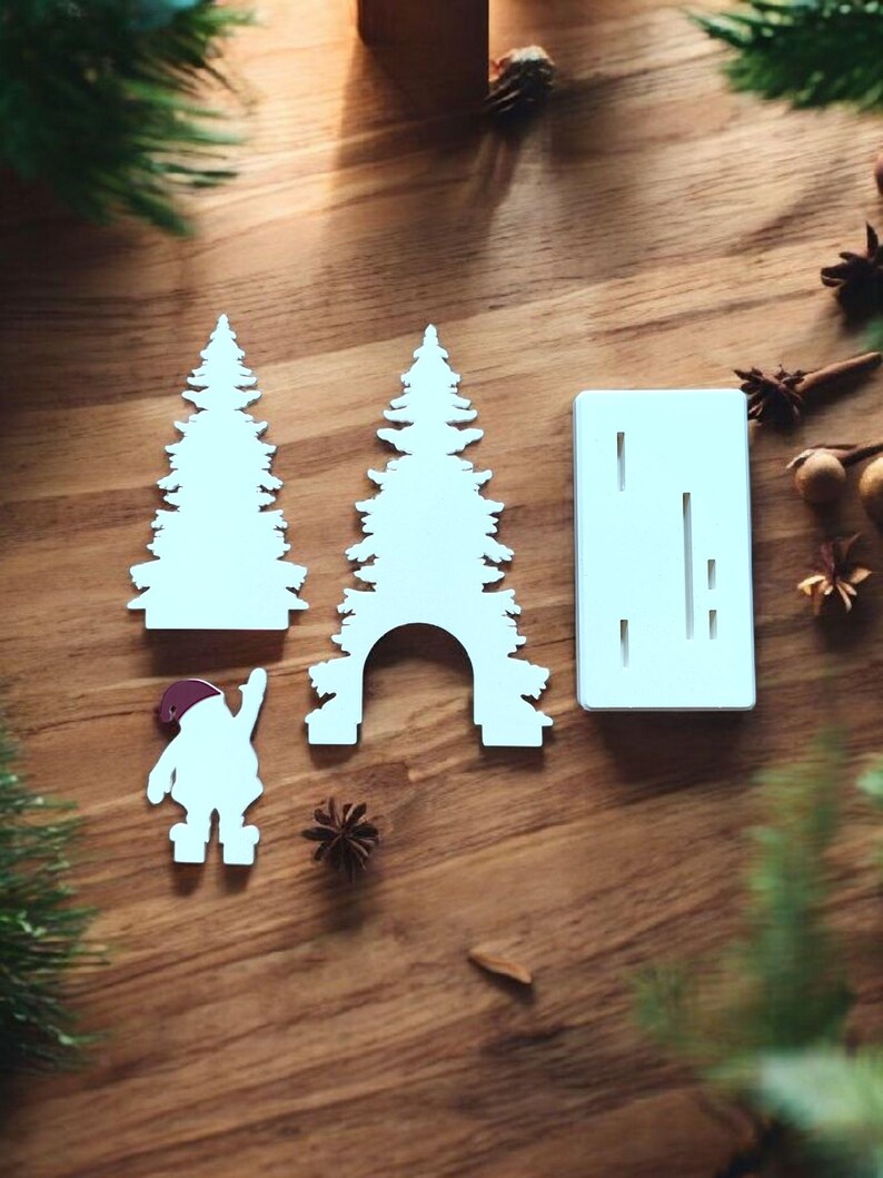 May include: White Christmas-themed cutouts on a wooden surface. The cutouts include two Christmas trees, a Santa Claus figure, and a rectangular piece with vertical slits. The Santa figure is wearing a red hat. The scene is decorated with pine needles and star anise.