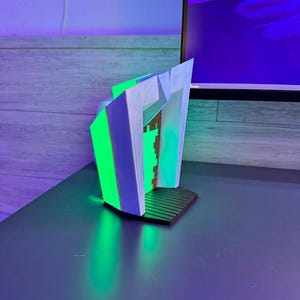 May include: A modern, abstract bookend design. The bookend features a geometric shape with white, green, and blue accents. The base is black, and the overall design is illuminated with green light, creating a futuristic aesthetic.