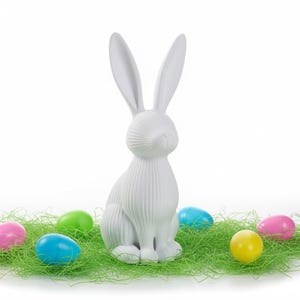 May include: A white ceramic Easter bunny figurine with long ears, sitting on green decorative grass. Colorful Easter eggs in pink, blue, green, and yellow surround the bunny, creating a festive spring scene.
