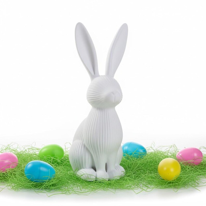 May include: A white ceramic Easter bunny figurine with long ears, sitting on green decorative grass. Colorful Easter eggs in pink, blue, green, and yellow surround the bunny, creating a festive spring scene.