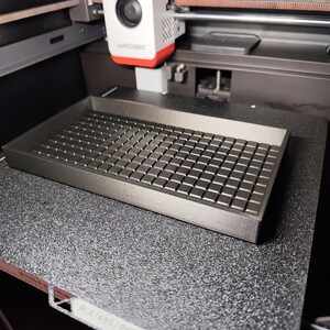 Op de afbeelding: Een donkergrijze 3D-geprinte tray met een rasterpatroon, geplaatst op een zwart oppervlak met textuur. De tray is rechthoekig met opstaande randen en een reeks kleine, vierkante blokken die het raster vormen. Een 3D-printerkop is ook zichtbaar.
