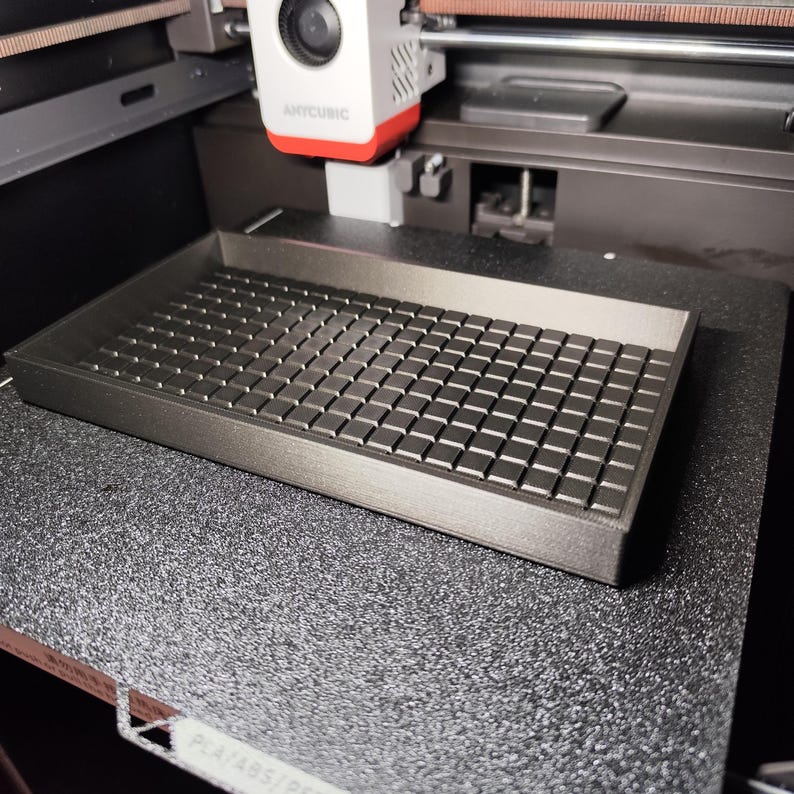 Op de afbeelding: Een donkergrijze 3D-geprinte tray met een rasterpatroon, geplaatst op een zwart oppervlak met textuur. De tray is rechthoekig met opstaande randen en een reeks kleine, vierkante blokken die het raster vormen. Een 3D-printerkop is ook zichtbaar.