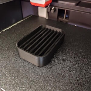 May include: A black, rectangular soap dish with a slatted design. The dish is made of a matte black material and sits on a textured surface. The dish has a raised edge and is designed to hold a bar of soap.