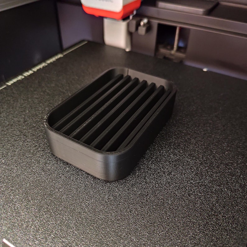 May include: A black, rectangular soap dish with a slatted design. The dish is made of a matte black material and sits on a textured surface. The dish has a raised edge and is designed to hold a bar of soap.
