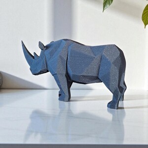 May include: A dark blue geometric rhino sculpture. The rhino is standing on a white surface. The sculpture has a textured surface and is made of a solid material. The rhino has a large horn and is a decorative item.