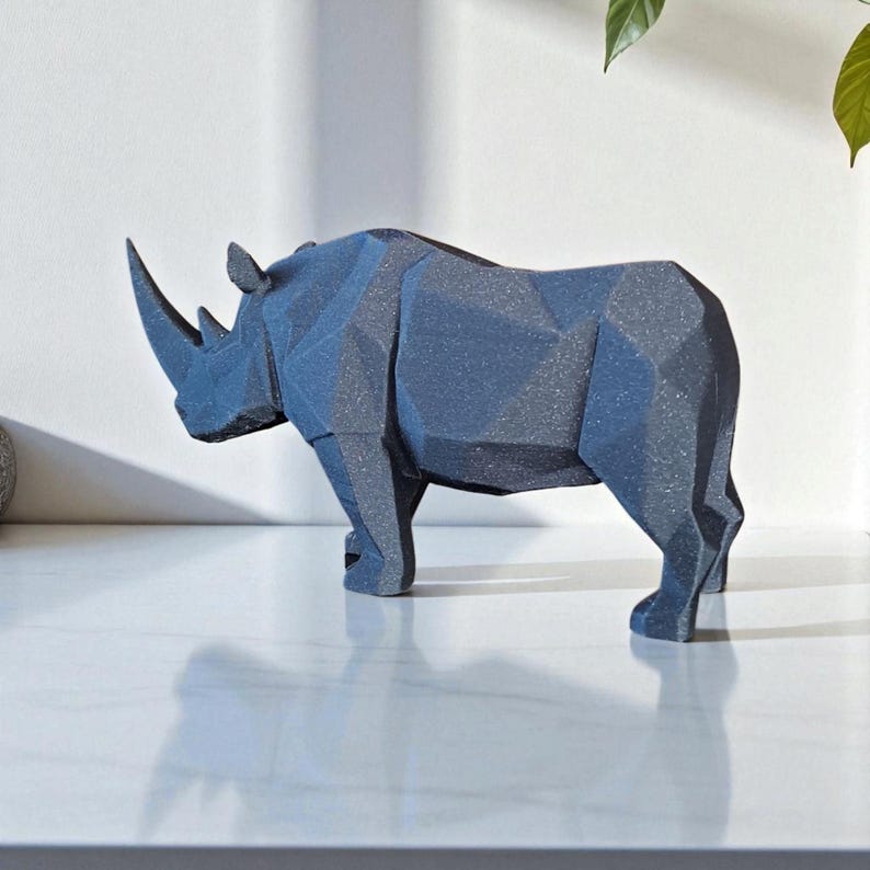 May include: A dark blue geometric rhino sculpture. The rhino is standing on a white surface. The sculpture has a textured surface and is made of a solid material. The rhino has a large horn and is a decorative item.