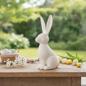 May include: A white ceramic Easter bunny figurine with long ears and a textured body sits on a wooden table. A small basket with pastel-colored eggs and spring flowers are also on the table, with a blurred green background.