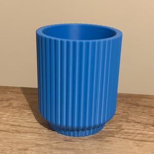 May include: A vibrant blue cylindrical planter with vertical ribbed detailing. The planter has a slightly flared base and a smooth interior. The color is a bright, solid blue, suitable for indoor or outdoor use.