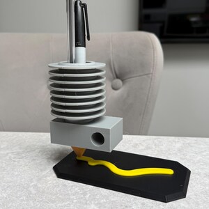 May include: A unique desk organizer with a black base and a gray pen holder. The pen holder has a cylindrical design with multiple tiers. A yellow line extends from the pen holder onto the base. A pen and a marker are in the holder.