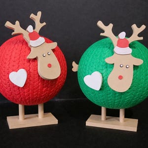 May include: Two Christmas reindeer decorations. One is red, the other green, each with a wooden reindeer head wearing a Santa hat. A white heart is on each sphere. The reindeer are on wooden bases.