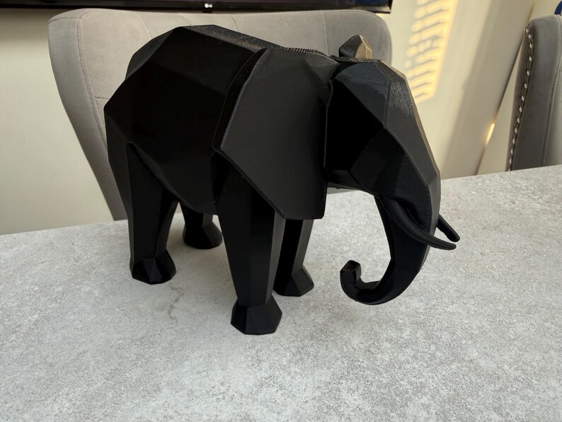 Op de afbeelding: Een zwarte geometrische olifant sculptuur. Het decoratieve stuk heeft een polygonale vormgeving met zichtbare randen en een omhoog gekrulde slurf. De olifant staat op vier poten en heeft grote oren. De sculptuur is gemaakt van een effen, matzwart materiaal.