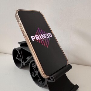 May include: Black phone stand with a curved base and a cradle to hold a phone. The phone displays the text "PRIM3D" in white and pink. The stand is designed to hold a phone at an angle.