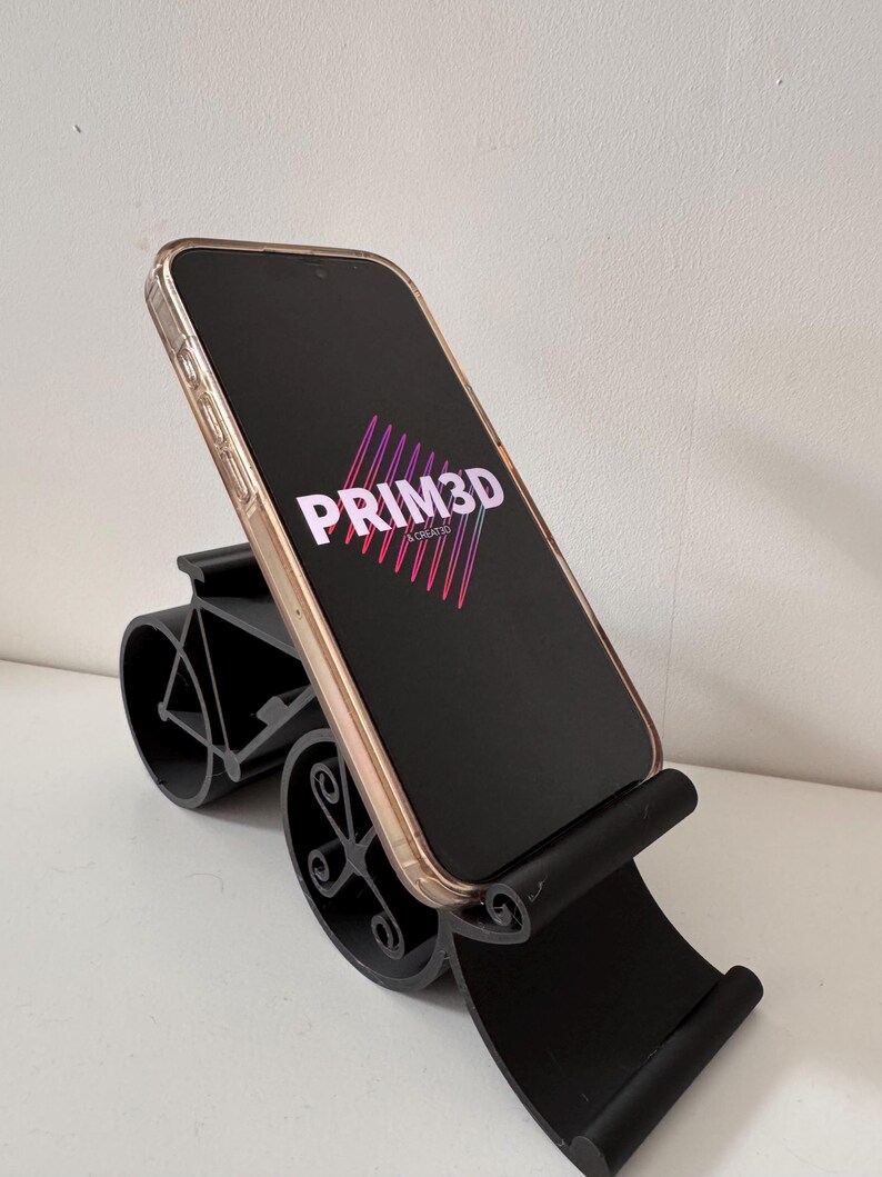 May include: Black phone stand with a curved base and a cradle to hold a phone. The phone displays the text "PRIM3D" in white and pink. The stand is designed to hold a phone at an angle.