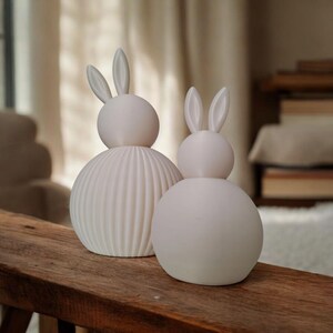 May include: Two white ceramic bunny figurines. One has a ribbed body, the other a smooth body. Both have rounded heads and long ears. They are displayed on a wooden surface, suggesting a decorative or Easter-themed item.