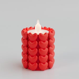May include: A red candle holder decorated with heart-shaped designs. The candle holder has a white, glowing LED light in the center, resembling a candle flame. The design is suitable for Valentine's Day or other romantic occasions.