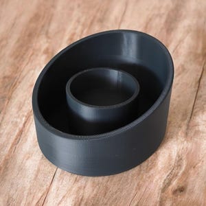 May include: A dark gray, 3D-printed candle holder with a circular inner well and an oval-shaped outer ring. The candle holder is made of a matte material and sits on a wooden surface.