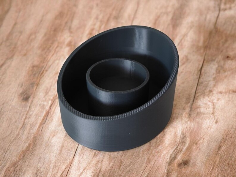 May include: A dark gray, 3D-printed candle holder with a circular inner well and an oval-shaped outer ring. The candle holder is made of a matte material and sits on a wooden surface.