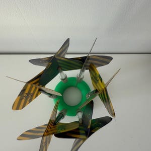 May include: A decorative object with a green circular base and six blades. The blades display a yellow and black striped pattern with green detailing. The object is set against a white background.