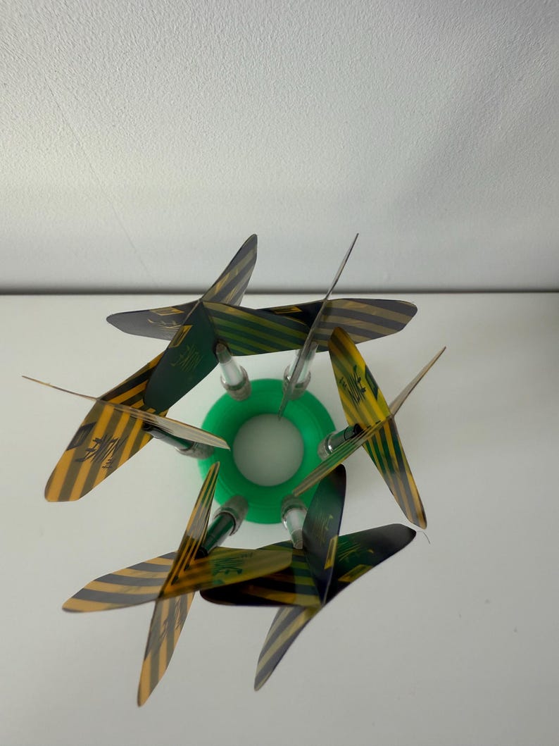 May include: A decorative object with a green circular base and six blades. The blades display a yellow and black striped pattern with green detailing. The object is set against a white background.