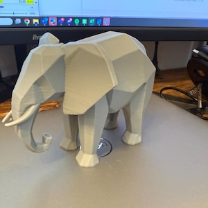 Op de afbeelding: Een lichtgrijze, geometrische olifantenfiguur. De olifant heeft een facetontwerp, met zichtbare randen en vlakken, waardoor hij een moderne, hoekige uitstraling heeft. De slurf is gekruld en de slagtanden zijn zichtbaar. De figuur is een decoratief item.