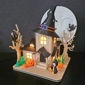 May include: A Halloween-themed decorative house with a witch's hat, pumpkins, and a full moon backdrop. The house has lit windows and a small figure holding a jack-o'-lantern. The color palette includes black, orange, beige, and green.