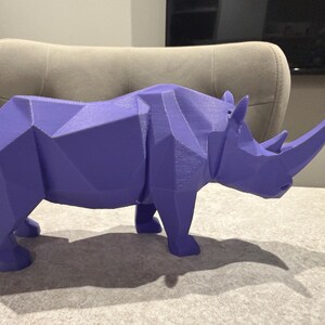 May include: A geometric, purple rhinoceros sculpture. The rhino is depicted in a low-poly style, with many flat, angled surfaces. The sculpture has a large horn and is standing on a table. The rhino's shadow is visible on the table.