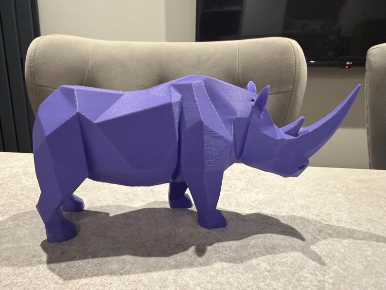May include: A geometric, purple rhinoceros sculpture. The rhino is depicted in a low-poly style, with many flat, angled surfaces. The sculpture has a large horn and is standing on a table. The rhino's shadow is visible on the table.