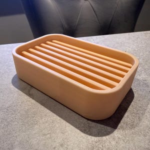 May include: A rectangular, peach-colored soap dish with a slatted top. The dish has rounded corners and is made of a solid material. The design allows for water drainage, making it a practical bathroom accessory.