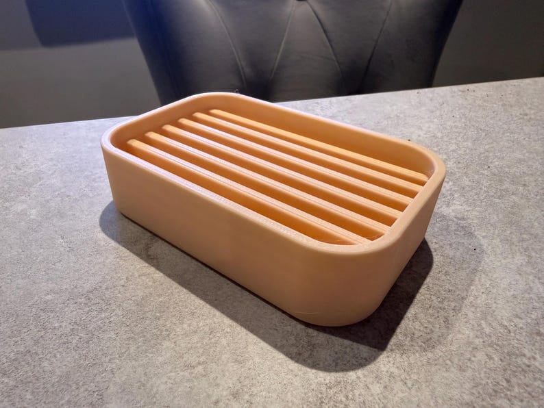 May include: A rectangular, peach-colored soap dish with a slatted top. The dish has rounded corners and is made of a solid material. The design allows for water drainage, making it a practical bathroom accessory.